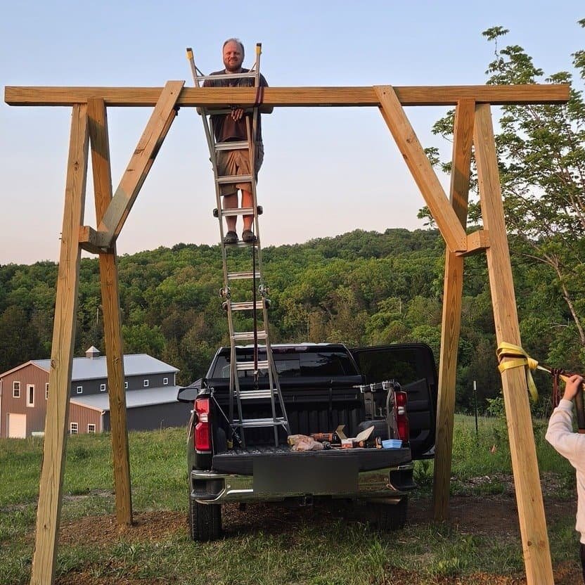 Wooden A-frame swing set frame standing in a field at golden hour, with a man on a ladder securing the top beam